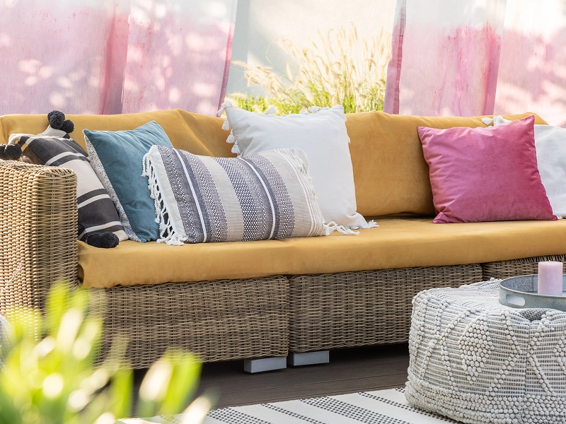 GettyImages-1143981413 Rattan outdoor sofa with mustard-yellow cushions and colorful throw pillows. Nearby, a round woven rug, a small circular table, and pink drapes in the background.